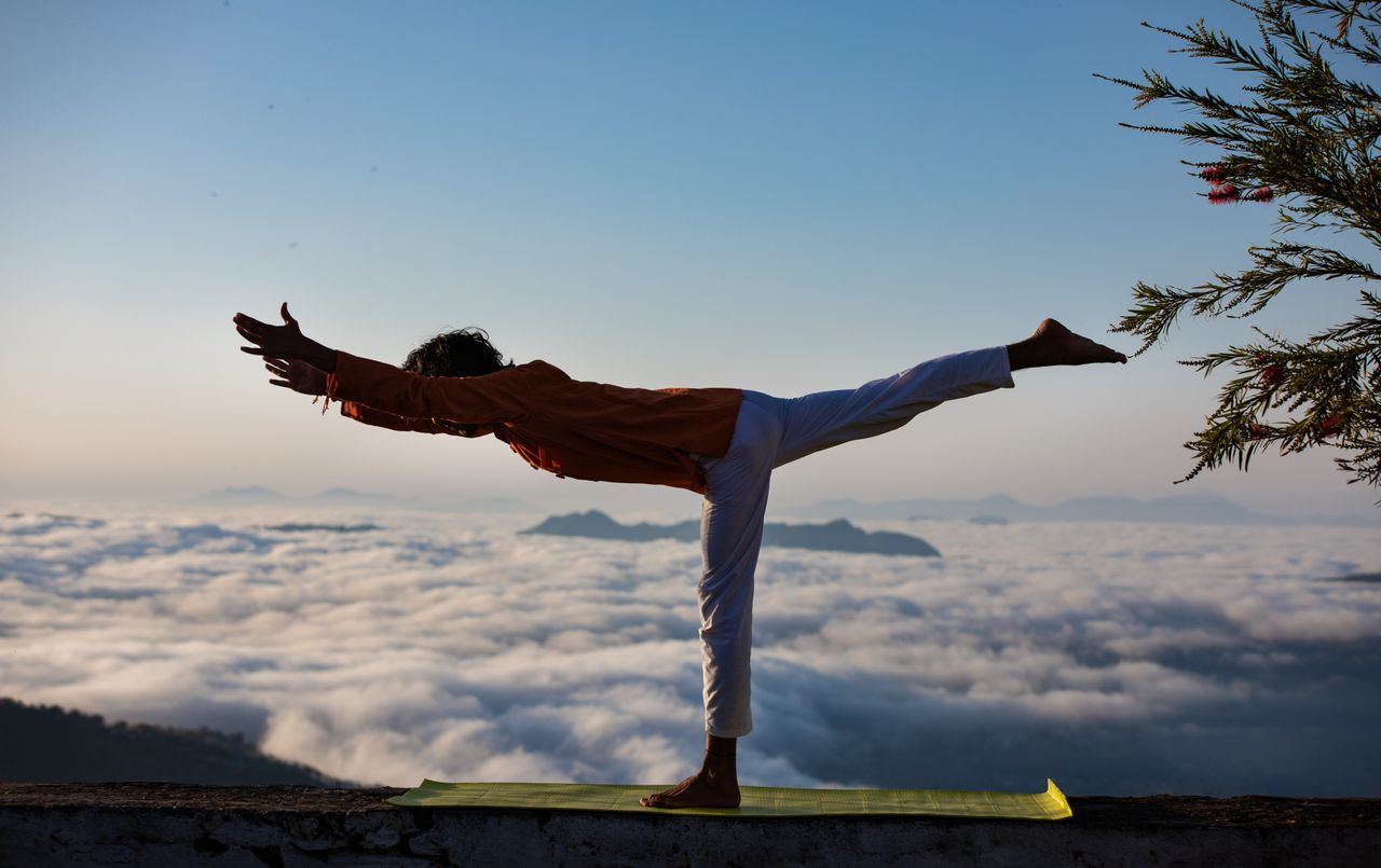 Home Person practicing yoga in warrior pose above a scenic cloudscape during sunrise.